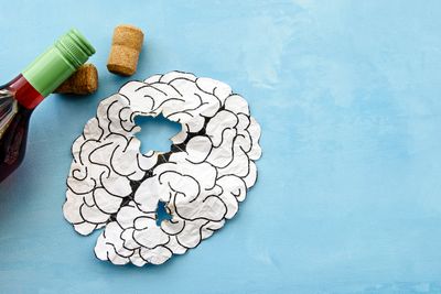 paper cutout of a brain with holes in it, next to a wine bottle and two corks on a blue background; illustration of alcohol-related brain damage