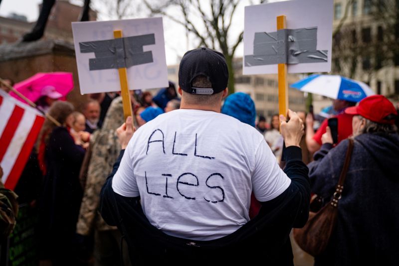 A photo of the back of a male protestor wearing a white T-shirt with "All Lies" written on the back. The man is wearing a black cap and is among a group of other protestors holding signs. We cannot see what is written on the signs are they are facing the other direction. 