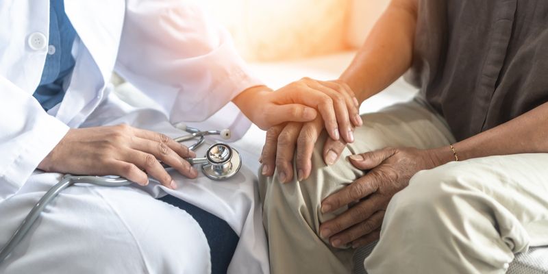 A close up photo of a female doctor holding the hand of an older man as they sit next to one another. The photo is cropped so that it is framing their laps, where we see the older man resting his hands on his leg as the doctor rests her left hand on his. The doctor is wearing a white coat and holding a stethoscope in her right hand.