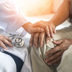 A close up photo of a female doctor holding the hand of an older man as they sit next to one another. The photo is cropped so that it is framing their laps, where we see the older man resting his hands on his leg as the doctor rests her left hand on his. The doctor is wearing a white coat and holding a stethoscope in her right hand.