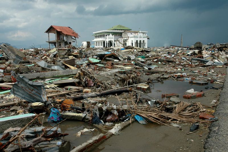 A photo of Banda Aceh, Aceh, in Indonesia after the 2004 tsunami. The photo shows the wreckage of various broken buildings, wood and metal debris piled on top of itself over some water. In the distance, one large white building is still standing amongst the destruction. The surrounding sky is grey and moody.