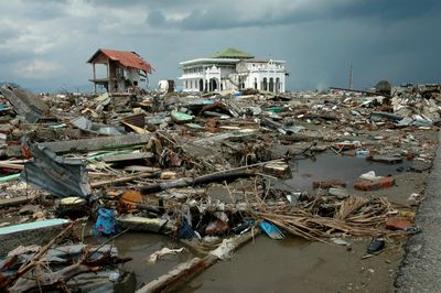 A photo of Banda Aceh, Aceh, in Indonesia after the 2004 tsunami. The photo shows the wreckage of various broken buildings, wood and metal debris piled on top of itself over some water. In the distance, one large white building is still standing amongst the destruction. The surrounding sky is grey and moody.