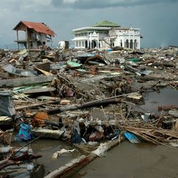 A photo of Banda Aceh, Aceh, in Indonesia after the 2004 tsunami. The photo shows the wreckage of various broken buildings, wood and metal debris piled on top of itself over some water. In the distance, one large white building is still standing amongst the destruction. The surrounding sky is grey and moody.