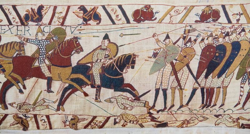 A scene from the Bayeux tapestry showing the invaders on horseback charging into a line of soldiers with spears and shields. Under there are some fallen soldiers under the approaching horses and arrows and spears are seen in the air. The margins above and below the image contain decorations, griffin like creatures and some additional fallen soldiers.  