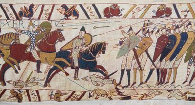 A scene from the Bayeux tapestry showing the invaders on horseback charging into a line of soldiers with spears and shields. Under there are some fallen soldiers under the approaching horses and arrows and spears are seen in the air. The margins above and below the image contain decorations, griffin like creatures and some additional fallen soldiers.  