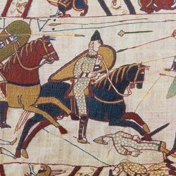 A scene from the Bayeux tapestry showing the invaders on horseback charging into a line of soldiers with spears and shields. Under there are some fallen soldiers under the approaching horses and arrows and spears are seen in the air. The margins above and below the image contain decorations, griffin like creatures and some additional fallen soldiers.  