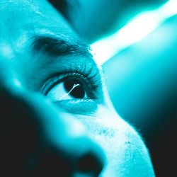 A man's face and eye upclose, under a bright blue teal light.