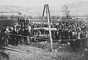 A photo showing the supposed Cardiff giant being lifted from the ground. The photo is black and white and grainy. It shows a dozens of people gathered around a scaffold that appears to be raising the giant out of the ground. The giant is laying horizonal in the middle of the shot and the foreground shows open grass and then a rope fence in front of the gathered people. A photo showing the supposed Cardiff giant being lifted from the ground. The photo is black and white and grainy. It shows a dozens of people gathered around a scaffold that appears to be raising the giant out of the ground. The giant is laying horizonal in the middle of the shot and the foreground shows open grass and then a rope fence in front of the gathered people.