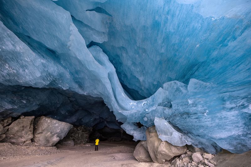 Person in yellow jacket stands inside a large ice cave with textured blue walls and rocky ground; Swiss Alps, Moteratsch Glacier