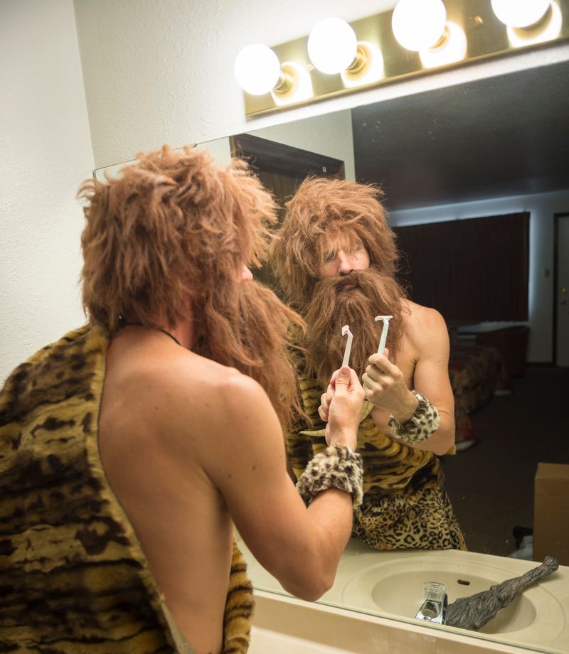 model of a stereotypical caveman wearing animal furs standing at a bathroom sink in the mirror, looking at a safety razor