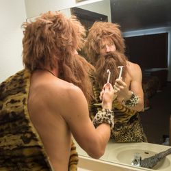 model of a stereotypical caveman wearing animal furs standing at a bathroom sink in the mirror, looking at a safety razor