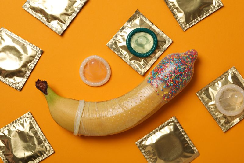 condom on a banana with sprinkles in the end of the condom; the banana is on an orange background and surrounded by a selection of wrapped and unwrapped condoms