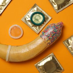 condom on a banana with sprinkles in the end of the condom; the banana is on an orange background and surrounded by a selection of wrapped and unwrapped condoms