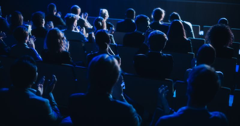 dark conference auditorium with lots of people sitting watching a lecture; they are viewed from behind so you can't see their faces