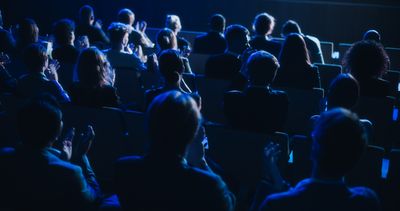 dark conference auditorium with lots of people sitting watching a lecture; they are viewed from behind so you can't see their faces