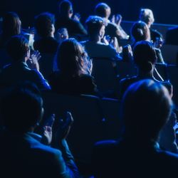 dark conference auditorium with lots of people sitting watching a lecture; they are viewed from behind so you can't see their faces