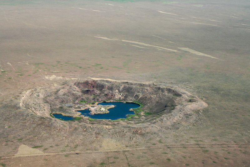 A photo taken from the air showing Lake Chagan in what was the Semipalatinsk Nuclear Test Site. The crater is located in a spars, almost desert like landscape and its quite round in shape. At its center is a horse-shoe shaped body of rich blue water.