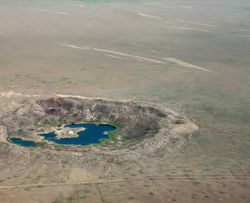 A photo taken from the air showing Lake Chagan in what was the Semipalatinsk Nuclear Test Site. The crater is located in a spars, almost desert like landscape and its quite round in shape. At its center is a horse-shoe shaped body of rich blue water.
