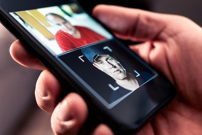 A photo showing someone's hand holding a smart phone. The phone's screen has two images on it, one above the other. In the top one, a man with a red t-shirt appears to be facing the screen, while the bottom image shows a close-up of a different man who is wearing a black cap and a black t-shirt. The second man has digital lines over the features of his face.