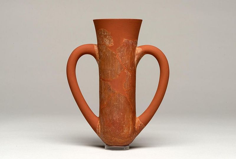A photo of a depas goblet, showing its characteristic narrow body with a wider top. The two handles curve from its sides, forming a kind of heart shape. The vessel is made from a red clay. 