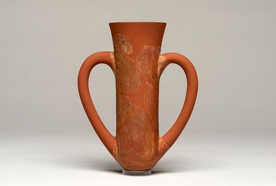 A photo of a depas goblet, showing its characteristic narrow body with a wider top. The two handles curve from its sides, forming a kind of heart shape. The vessel is made from a red clay. 