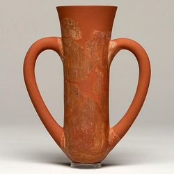 A photo of a depas goblet, showing its characteristic narrow body with a wider top. The two handles curve from its sides, forming a kind of heart shape. The vessel is made from a red clay. 