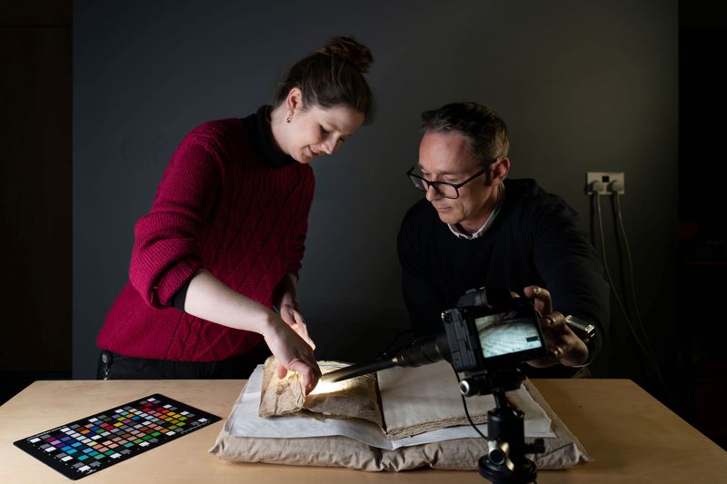 The photo shows two researchers, a woman wearing a red sweater with the sleeves rolled up, and a man wearing a black sweater and glasses, examining a manuscript laid out on a cushion support. The woman is holding a page of the manuscript while a camera with a long lens photographs it. Next to the manuscript is a colour chart resting on the table.
