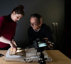 The photo shows two researchers, a woman wearing a red sweater with the sleeves rolled up, and a man wearing a black sweater and glasses, examining a manuscript laid out on a cushion support. The woman is holding a page of the manuscript while a camera with a long lens photographs it. Next to the manuscript is a colour chart resting on the table.