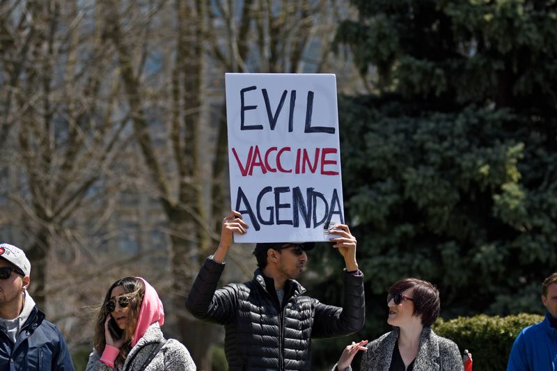 A man in a black puffer jacket and sunglasses holds up a large piece of card saying "Evil Vaccine Agenda" while other protestors stand around him. 