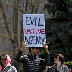 A man in a black puffer jacket and sunglasses holds up a large piece of card saying "Evil Vaccine Agenda" while other protestors stand around him. 