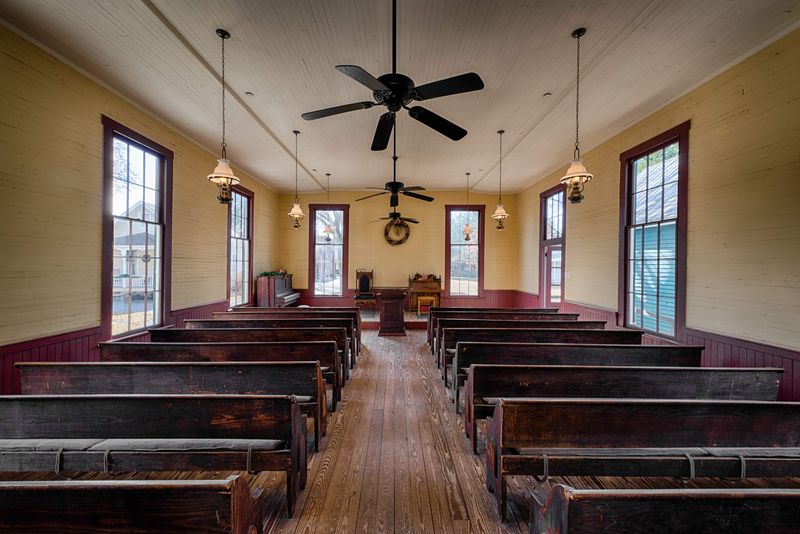 A photo taken within a small American church. The camera is positioned within the center of the nave and shows seven rows of pews either side. There are also size large windows equally spaces at the front of the church. The image has low hanging lamps suspended from the ceiling, and there are two large ceiling fans in view. 