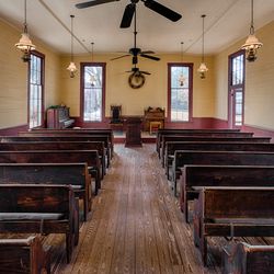 A photo taken within a small American church. The camera is positioned within the center of the nave and shows seven rows of pews either side. There are also size large windows equally spaces at the front of the church. The image has low hanging lamps suspended from the ceiling, and there are two large ceiling fans in view. 