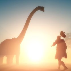 Silloute of caveman people walking towards a dinosaur in the sunset.