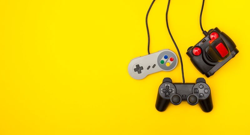 A photo showing three retro gaming control pads hanging against an yellow background by their cables. All three controllers are positioned towards the right hand side of the image. The first is a super Nintendo control, next is a playstation 2 controller positioned a little lower, and then there is a classic Atari joystick controller.