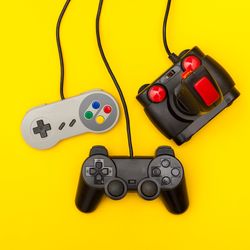 A photo showing three retro gaming control pads hanging against an yellow background by their cables. All three controllers are positioned towards the right hand side of the image. The first is a super Nintendo control, next is a playstation 2 controller positioned a little lower, and then there is a classic Atari joystick controller.