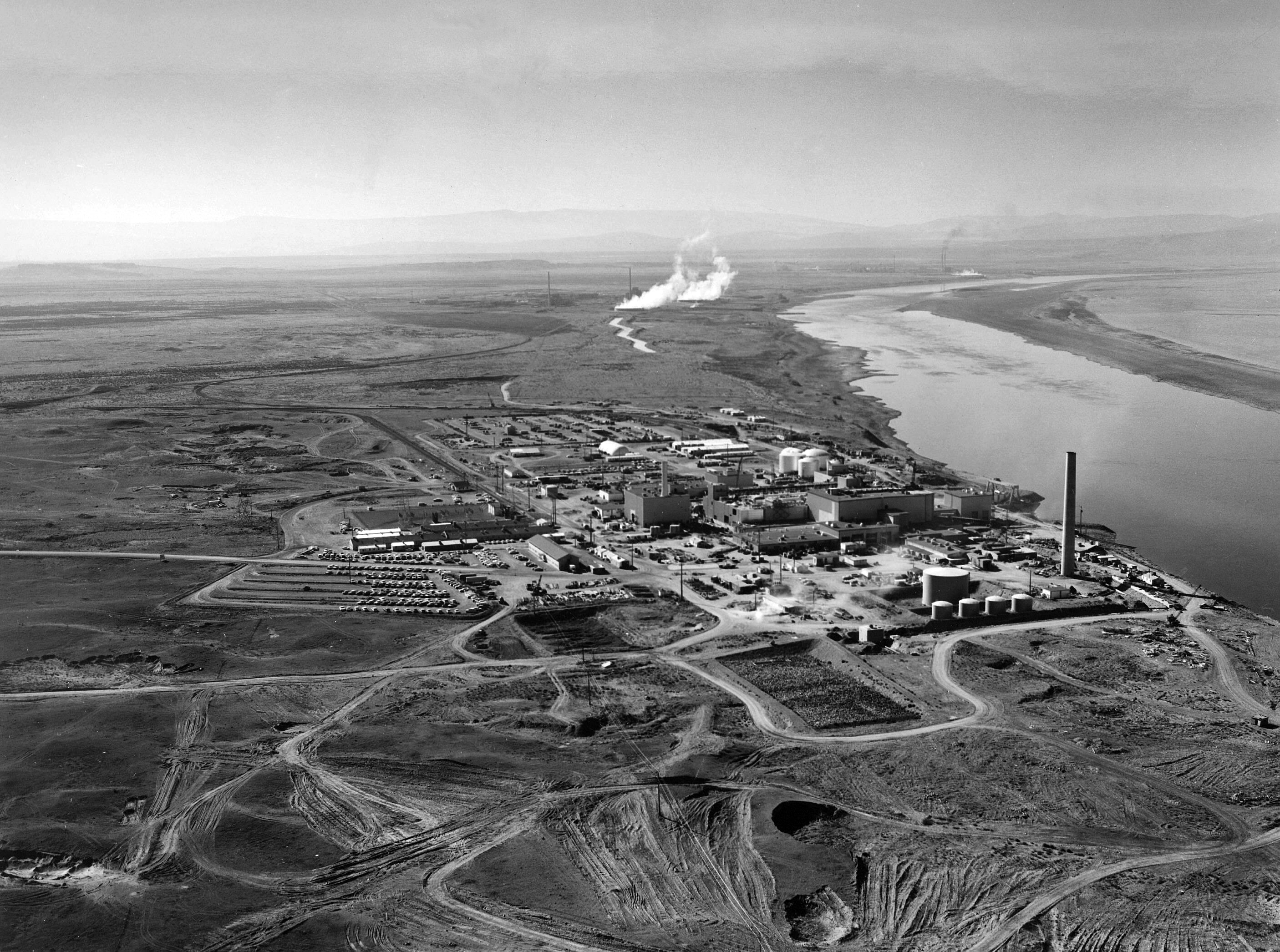 A black and white aerial photo showing the reactor complex by the Columbia River. The land surrounding the site has been dug up and is showing the muddy top soil. In the distance, near the water's edge are plumes of steam rising from buildings fading into the distance. The land beyond the complex is largely rolling fields leading to mountains that are just about visible. A black and white aerial photo showing the reactor complex by the Columbia River. The land surrounding the site has been dug up and is showing the muddy top soil. In the distance, near the water's edge are plumes of steam rising from buildings fading into the distance. The land beyond the complex is largely rolling fields leading to mountains that are just about visible.