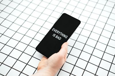 A photo of a person's hand holding a smart phone with the words "Everything is bad" on the screen. The and hand is set against a black and white grid background. 
