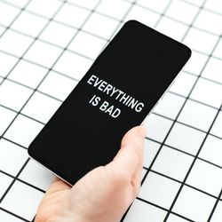 A photo of a person's hand holding a smart phone with the words "Everything is bad" on the screen. The and hand is set against a black and white grid background. 