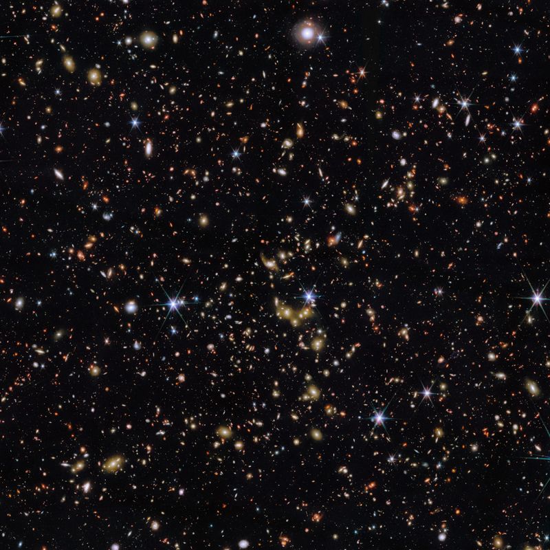 An area of deep space with thousands of galaxies in various shapes and sizes on a black background. Most are circles or ovals, with a few spirals. More distant galaxies are redder in colour and smaller, down to being mere dots, while closer galaxies are a bit larger and white or blueish. A few gold-coloured galaxies are bunched closely together in the centre. Bright stars surrounded by spikes lie in our galaxy.]
