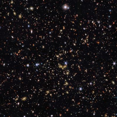 An area of deep space with thousands of galaxies in various shapes and sizes on a black background. Most are circles or ovals, with a few spirals. More distant galaxies are redder in colour and smaller, down to being mere dots, while closer galaxies are a bit larger and white or blueish. A few gold-coloured galaxies are bunched closely together in the centre. Bright stars surrounded by spikes lie in our galaxy.]