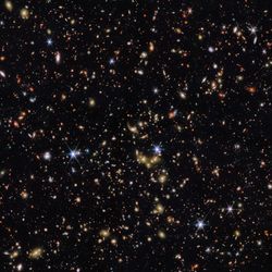 An area of deep space with thousands of galaxies in various shapes and sizes on a black background. Most are circles or ovals, with a few spirals. More distant galaxies are redder in colour and smaller, down to being mere dots, while closer galaxies are a bit larger and white or blueish. A few gold-coloured galaxies are bunched closely together in the centre. Bright stars surrounded by spikes lie in our galaxy.]