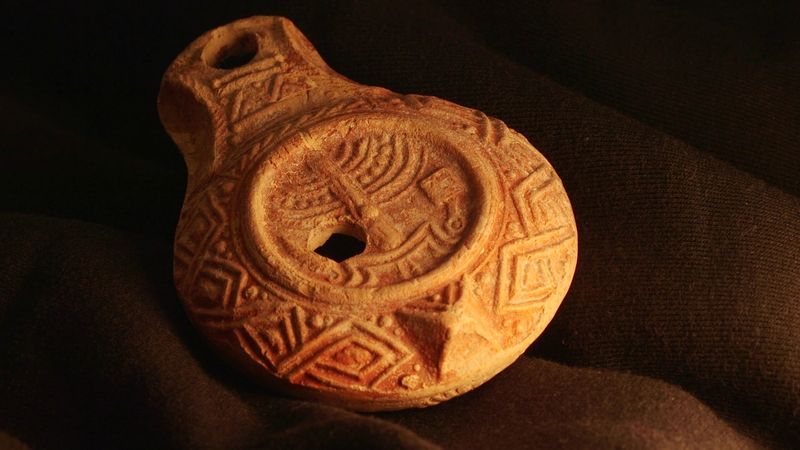 The photo shows the lamp lit by a soft orange light. The lamp is roughly tear-drop shaped and made of ceramic. It's surface has intricate decorations across it and, in the center, is the symbol of the menorah.