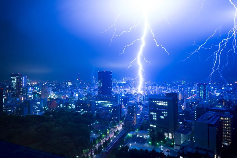 A photo taken from a high elevation at night shows Tokyo as a massive lightning strikes the ground in the middle of the image. The lightning is bright white, illuminating the city and casting a blue color around it. There are smaller, thinner lines of lightning to the right. 