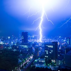 A photo taken from a high elevation at night shows Tokyo as a massive lightning strikes the ground in the middle of the image. The lightning is bright white, illuminating the city and casting a blue color around it. There are smaller, thinner lines of lightning to the right. 
