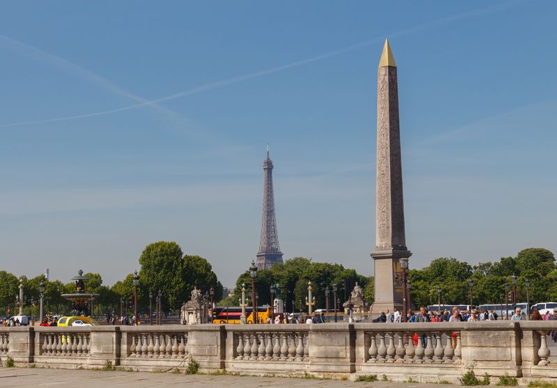 A photo showing the Luxor obelisk with the Eiffel Tower in the background. The obelisk has its characteristic golden pointed top and you can just make out some of the hieroglyphs on its side. The photo sits within a wider shot that also captures the busy tourists walking up the Place de la Concorde.