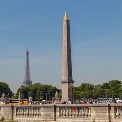 A photo showing the Luxor obelisk with the Eiffel Tower in the background. The obelisk has its characteristic golden pointed top and you can just make out some of the hieroglyphs on its side. The photo sits within a wider shot that also captures the busy tourists walking up the Place de la Concorde.