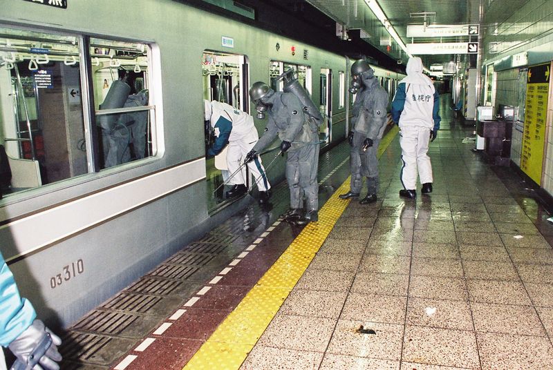 A photo showing emergency response agents in protective suits spraying down a train carriage on the Tokyo Underground. There are four workers in the shot approaching the carriage which is on the left of the shot. The one furthest away is in a white plastic full body suit and has his back to the camera. Next to him are two men in grey plastic suits and visible gas masks spraying the floor with water, while another man in a white plastic suit is leaning into the carriage. 