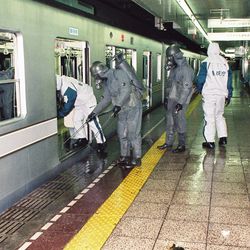 A photo showing emergency response agents in protective suits spraying down a train carriage on the Tokyo Underground. There are four workers in the shot approaching the carriage which is on the left of the shot. The one furthest away is in a white plastic full body suit and has his back to the camera. Next to him are two men in grey plastic suits and visible gas masks spraying the floor with water, while another man in a white plastic suit is leaning into the carriage. 
