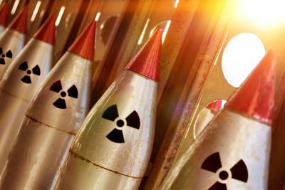 A photo showing a line of nuclear warheads attached to missiles. They are the pointed red tips on five weapons that have the radiation symbol just under them. The image shows the tops of these missiles lined up against a metal stand that has circular gaps in it which are letting in light.