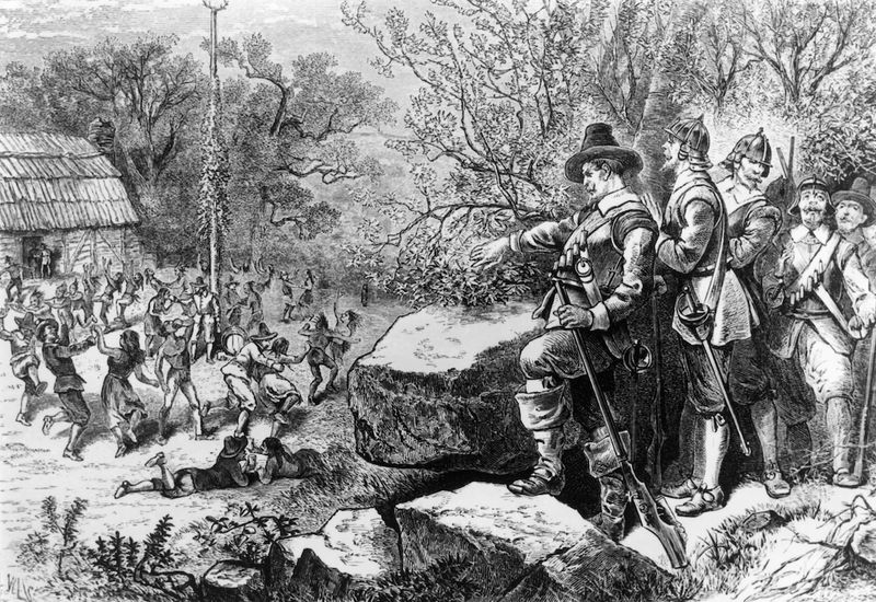 An 18th century engraving showing Puritans standing among some rocks looking down on settlers and Indigenous Americans dancing around a may-pole. The Puritans are all wearing armour as they are ready to clamp down on the spectacle below.  s 